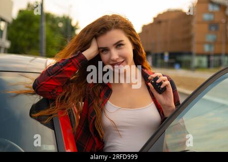 Con lucchetti rossi infuocati e un sorriso sorridente, la giovane donna che indossa una camicia a quadri casual posa accanto all'auto rossa e tiene le chiavi dell'auto in mano. Foto Stock