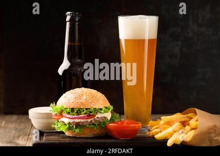 Hamburger, birra in bottiglia e in tazza di vetro, patatine fritte, salsa su sfondo scuro, concetto di fast food. Foto Stock