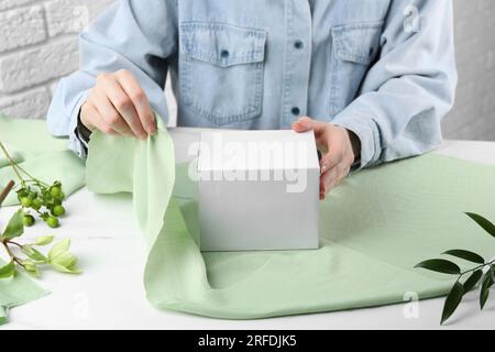 Tecnica Furoshiki. Donna che avvolge il regalo in tessuto verde al tavolo bianco, primo piano Foto Stock