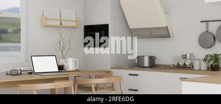 Mockup con schermo bianco per computer portatile, cuffie e arredamento su un tavolo da pranzo in legno in una cucina bianca minimalista con moderni elettrodomestici da cucina. Lavoro domestico Foto Stock