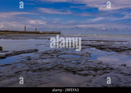 Whitby East Pier con la bassa marea, vista dalle rocce di scisto nella spiaggia orientale. Il cielo blu si riflette nelle piscine rocciose sulla spiaggia. Foto Stock