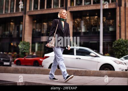 Bella giovane donna elegante che cammina lungo la strada della città sullo sfondo di un edificio, indossa abiti alla moda jeans, blazer nero, borsa e snea Foto Stock