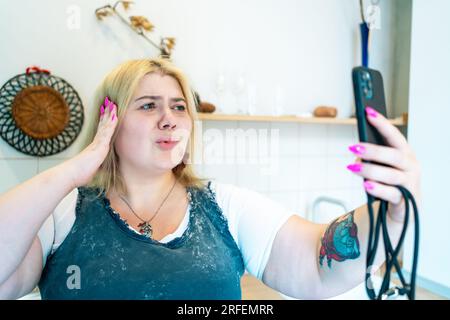 Giovane donna bionda positiva Plus size che effettua videochiamate o selfie con il cellulare in cucina a casa. Foto Stock