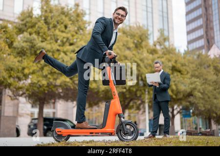 Affari pazzi, veloci e facili. Un uomo d'affari eccitato e divertente su uno scooter elettrico per le strade cittadine. Impiegato uomo d'affari avvocato aziendale in suit riding e Foto Stock