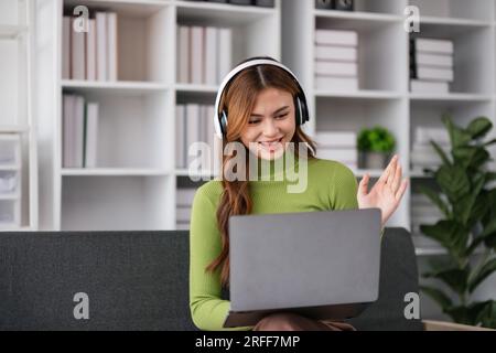 Bella giovane donna asiatica indossa le cuffie sorridenti dai il benvenuto tramite chat videochiamate sul computer portatile, ragazza rilassati goditi la musica online Foto Stock