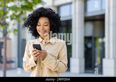 Giovane donna afroamericana con il telefono sorridente, donna d'affari fuori dall'edificio dell'ufficio utilizza l'app per smartphone, digita messaggi e naviga nelle pagine Internet. Foto Stock