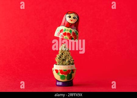 Una matryoshka di legno si erge su uno sfondo rosso, in esso è un bocciolo secco di marijuana medica. Trattamento alternativo con cannabis medica Foto Stock