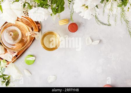 Teiera alle erbe, tazza, dessert con macaron, sfondo di stile di vita con fiori bianchi. Estetica femminile, tempo di meditazione per te stesso, concetto di vita lenta Foto Stock