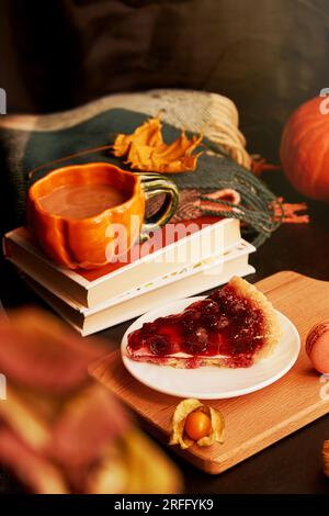 Colazione estetica con una tazza di caffè a forma di zucca, torta di ciliegie, macaron, candela e ciliegia invernale. Primo piano sfocato. Hygge accogliente casa. Foto Stock