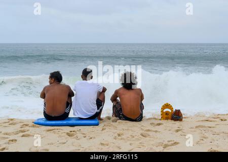 Niteroi, Brasile, vista posteriore di tre uomini brasiliani seduti sulla spiaggia di sabbia. Stanno guardando le onde infrangersi violentemente durante la stagione invernale. Foto Stock