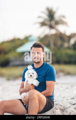 Giovane con il suo piccolo cane bianco maltese sulla spiaggia al tramonto Foto Stock