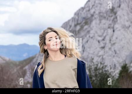 Mountain Beauty Unleashed: L'affascinante Ritratto di una giovane donna bionda che abbraccia la maestà della natura Foto Stock
