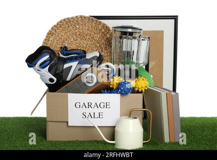 Scatola con cartello Garage sale e cose diverse su erba verde su sfondo bianco Foto Stock