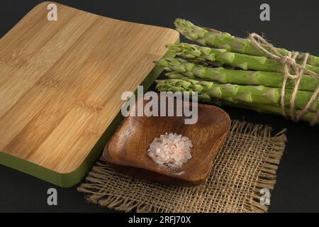 Mazzo di asparagi verdi legato con filo di iuta vicino al tagliere su sfondo nero con spazio copia. Cottura degli asparagi e concep di verdure primaverili Foto Stock