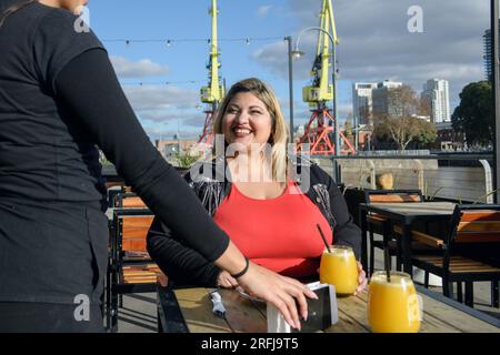 giovane donna latina bionda, sorridente seduta fuori dal ristorante al tramonto servita dalla cameriera, durante l'ora dello spuntino. spazio copia, restaura Foto Stock