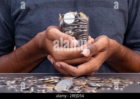 Un uomo in abiti tradizionali indiani tiene in mano una manciata di monete indiane. Le monete sono sparse su un tavolo di vetro davanti a lui Foto Stock