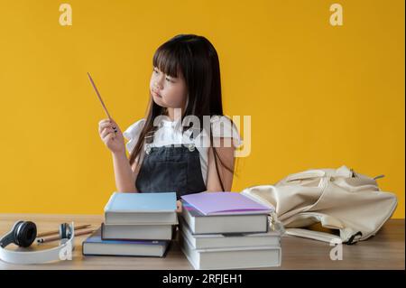 Un'adorabile giovane ragazza asiatica sta preparando la sua cancelleria per la sua giornata scolastica al suo tavolo su uno sfondo giallo isolato. Concetto di studentessa Foto Stock