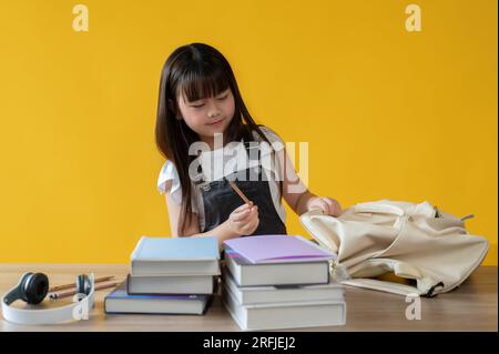 Un'adorabile giovane ragazza asiatica sta preparando la sua cancelleria o imballando la borsa della scuola per la sua giornata scolastica al suo tavolo contro uno zaino giallo isolato Foto Stock