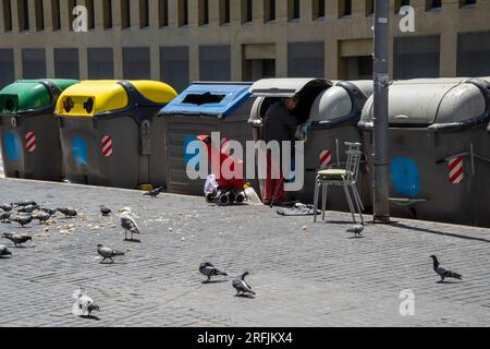 Senzatetto con un bastone in cerca di cibo in un contenitore di spazzatura in una strada di Barcellona con il suo carrello della spesa e le borse accanto, e molti piccioni Foto Stock