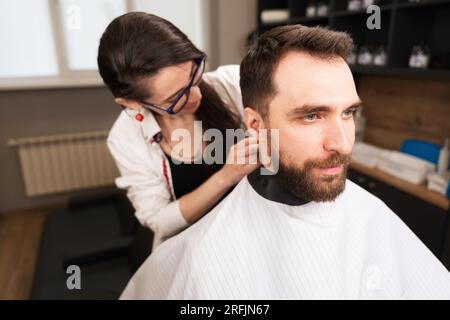 Una parrucchiere indossa un mantello protettivo a un uomo barbuto Foto Stock