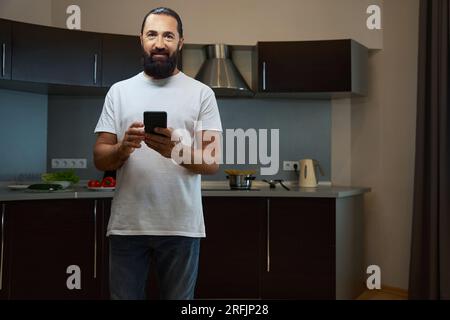 Bell'uomo è in piedi nella zona cucina e sta scrivendo su un telefono cellulare, sta indossando dei jeans comodi Foto Stock