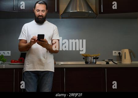 L'uomo barbuto sta in cucina e invia messaggi sul cellulare Foto Stock