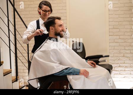 Bell'uomo siede in un barbiere su una sedia da barbiere, una donna gli fa un taglio di capelli con le forbici Foto Stock