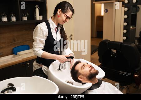 La femmina lava la testa dei clienti con acqua calda, maschio in un mantello protettivo Foto Stock