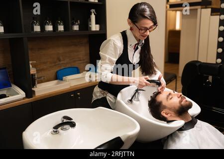 La giovane donna barbiera lava i capelli di un uomo, il cliente si trova in un lavandino speciale Foto Stock