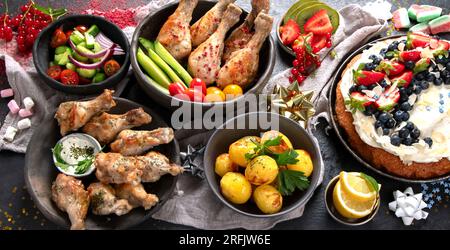 Cena con pollo, verdure, torta, insalata e frutta su sfondo scuro. Concetto di cibo estivo. Vista dall'alto. Foto Stock