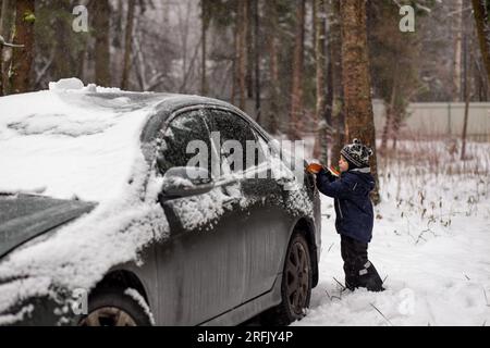 un bambino carino che aiuta a spazzolare la neve da un'auto durante la giornata invernale in campagna. Immagine con messa a fuoco selettiva su un ragazzo. Foto di alta qualità Foto Stock