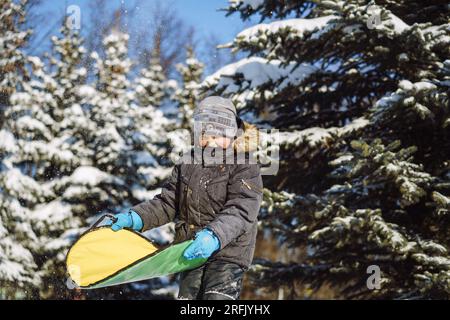 un simpatico ragazzo caucasico che tiene in mano un disco di neve per scivolare dalla collina nel parco invernale. Foto di alta qualità Foto Stock