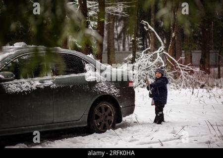 un bambino carino che aiuta a spazzolare la neve da un'auto durante la giornata invernale in campagna. Immagine con messa a fuoco selettiva su un ragazzo. Foto di alta qualità Foto Stock