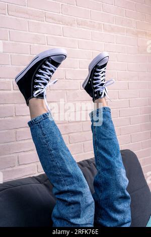 una ragazza adolescente, vestita di jeans blu e di nuove sneakers con allacciatura, è sdraiata sul divano con le gambe sollevate. Un concetto alla moda in stile casual Foto Stock