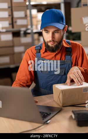 Responsabile del magazzino che utilizza un computer portatile per controllare il numero d'ordine su un pacco, preparando la scatola di cartone per la spedizione. Foto Stock