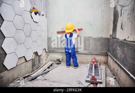 Bambino che controlla la superficie della parete con la livella a bolla d'aria nell'appartamento. Vista posteriore del bambino in tute da lavoro e casco da costruzione di sicurezza utilizzando lo strumento di livellamento durante il lavoro di ristrutturazione domestica. Foto Stock