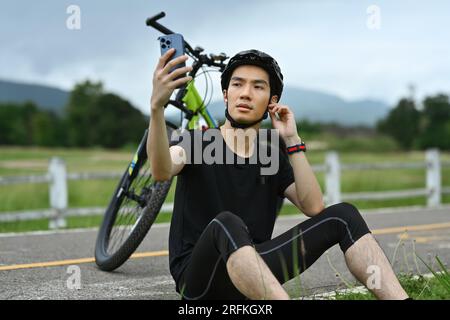 Ciclista maschio asiatico con casco nero riposa dopo aver pedalato in bicicletta e utilizzando lo smartphone. Foto Stock