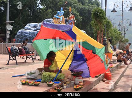 Una povera donna che vende oggetti puja a Triveni Ghat, Rishikesh, che tiene un ombrello per proteggersi dal sole. Rishikesh. Uttarakhand. India. Asia. Foto Stock