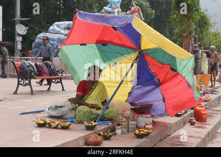 Una povera donna che vende oggetti puja a Triveni Ghat, Rishikesh, che tiene un ombrello per proteggersi dal sole. Rishikesh. Uttarakhand. India. Asia. Foto Stock