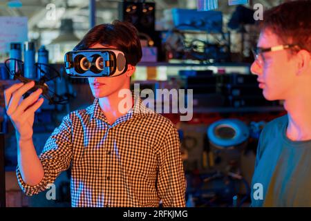 Adolescenti che fanno esperimenti di robotica in laboratorio. Ragazzo con visore VR in possesso di un piccolo drone. Illuminazione rossa e blu Foto Stock