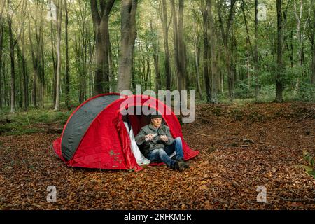 Uomo seduto davanti alla sua tenda nel mezzo di una foresta che versa un caffè da un thermos Foto Stock