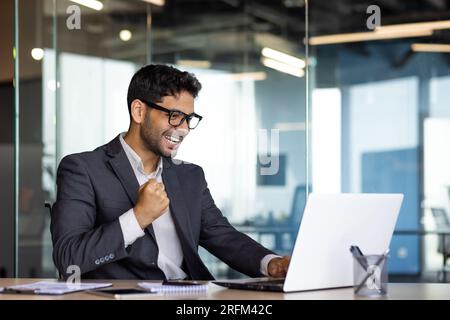 Giovane uomo d'affari arabo di successo sul posto di lavoro all'interno dell'ufficio, uomo in tuta aziendale con notebook che celebra la vittoria ha ricevuto una notifica online di vittoria, il vincitore del boss si rallegra e celebra il trionfo. Foto Stock