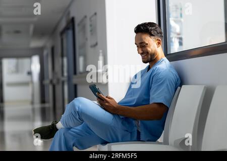 Medico maschio birazziale che indossa scrub utilizzando smartphone in corridoio all'ospedale Foto Stock