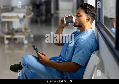 Medico maschio birazziale che indossa scrub bere caffè utilizzando smartphone in corridoio all'ospedale Foto Stock