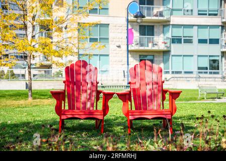 Due sedie rosse Adirondack su erba in un parco pubblico con condomini sullo sfondo in una soleggiata mattinata autunnale Foto Stock