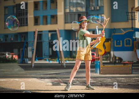 Una graziosa ragazza in età scolare soffia grandi bolle di sapone nel cortile di un edificio a più piani in estate. concetto estivo, giochi all'aperto Foto Stock