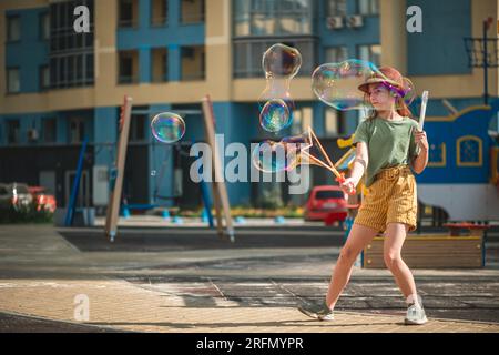 Una graziosa ragazza in età scolare soffia grandi bolle di sapone nel cortile di un edificio a più piani in estate. concetto estivo, giochi all'aperto Foto Stock