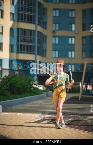 Una graziosa ragazza in età scolare soffia grandi bolle di sapone nel cortile di un edificio a più piani in estate. concetto estivo, giochi all'aperto Foto Stock