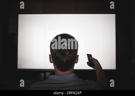 Uomo che guarda la televisione, accende il televisore a schermo piatto al plasma, punta il telecomando verso lo schermo TV vuoto sulla parete. Guy che cambia canale a casa, sul retro Foto Stock
