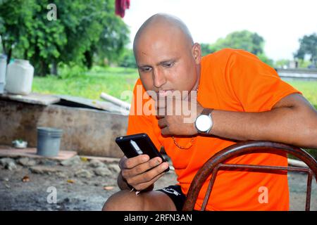 Simpatico modello calvo che indossa una t-shirt arancione, uomini allegri che guardano il telefono cellulare, lo sfondo della fattoria Foto Stock